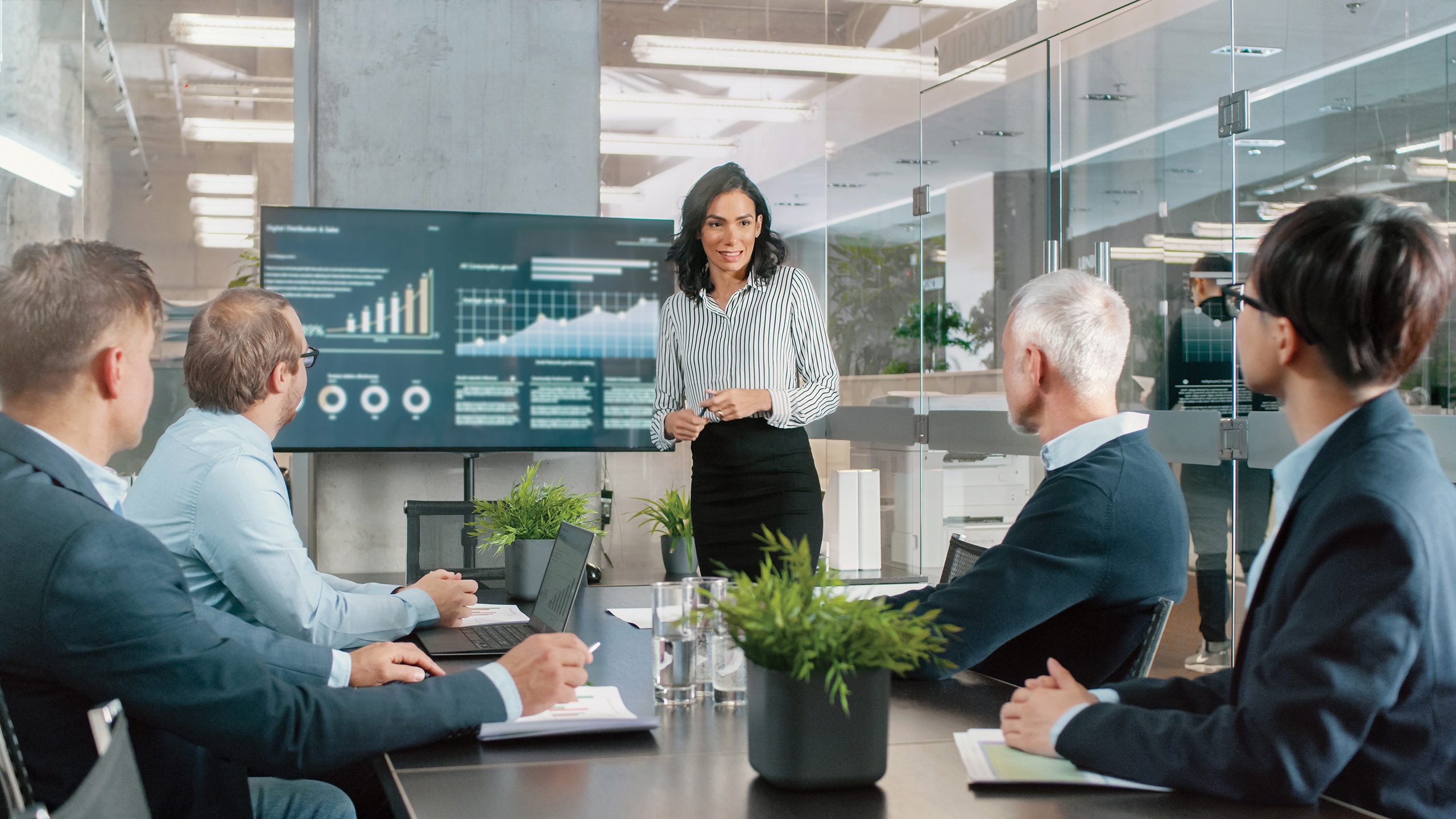 Beautiful Businesswoman Gives Report/ Presentation to Her Business Colleagues in the Conference Room, She Shows Graphics, Pie Charts and Company's Growth on the Wall TV.